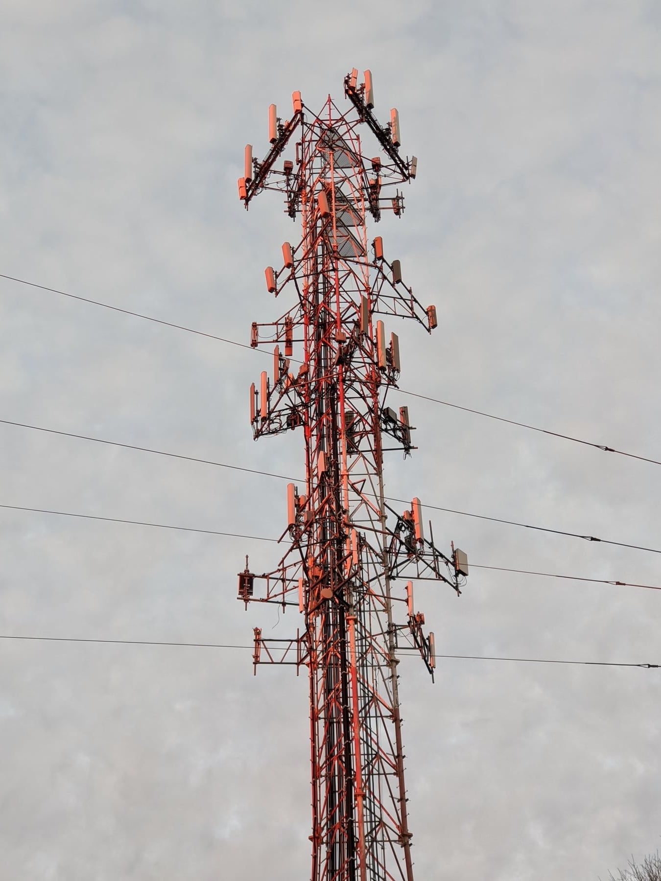 Red antenna scaffold tower rising against overcast grey sky, cross-braced steel and signal arrays